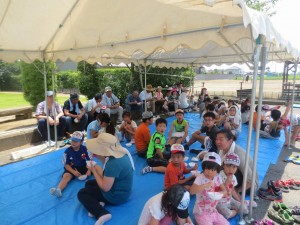 IMG_4428 写真は,7月26日(日) とよつまちづくりセンターの前です。子どもたちや地域の方々が,野菜の収穫を終え,バーベキューの準備として取れた野菜を切った後,かき氷を食べながら休憩しているところです。\